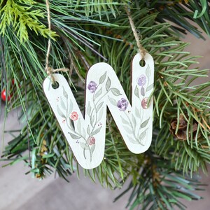 May include: A white wooden ornament shaped like the letter "W" with a floral design painted in shades of green, purple, and red. The ornament is hanging from a brown twine string.