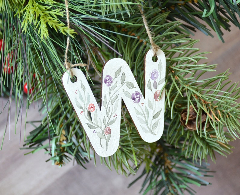 May include: A white wooden ornament shaped like the letter "W" with a floral design painted in shades of green, purple, and red. The ornament is hanging from a brown twine string.
