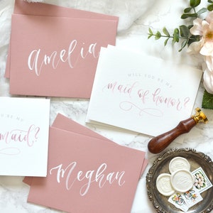 May include: Four cards with pink and white calligraphy. The cards say "Will you be my maid of honor?" and "Will you be my bridesmaid?" with the names Amelia and Meghan on the other two cards.