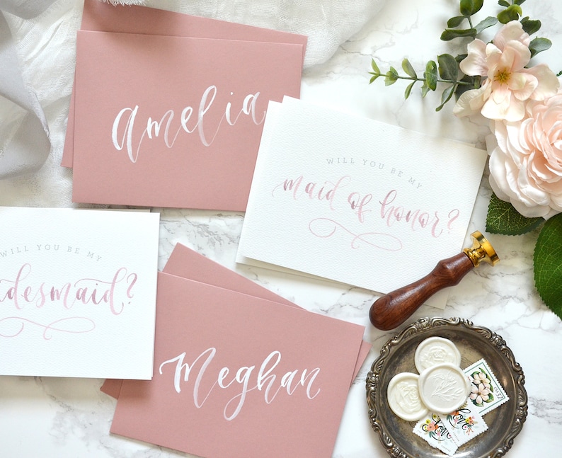 May include: Four cards with pink and white calligraphy. The cards say "Will you be my maid of honor?" and "Will you be my bridesmaid?" with the names Amelia and Meghan on the other two cards.