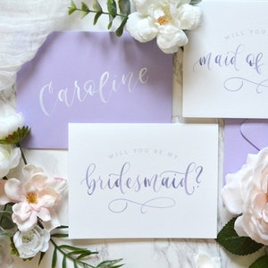 May include: Two purple envelopes with white calligraphy that reads "Caroline" and "Will you be my maid of honor?"  A white envelope with purple calligraphy that reads "Will you be my bridesmaid?"  The envelopes are surrounded by white and pink flowers.