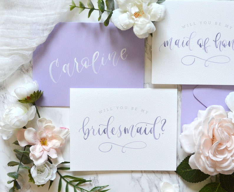 May include: Two purple envelopes with white calligraphy that reads "Caroline" and "Will you be my maid of honor?"  A white envelope with purple calligraphy that reads "Will you be my bridesmaid?"  The envelopes are surrounded by white and pink flowers.
