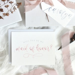 May include: A white card with the text "Will you be my maid of honor?" written in pink calligraphy. The card is surrounded by pink ribbon and a white envelope.
