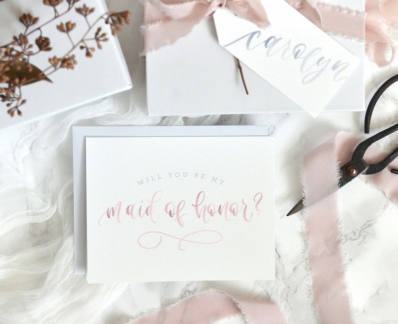 May include: A white card with the text "Will you be my maid of honor?" written in pink calligraphy. The card is surrounded by pink ribbon and a white envelope.