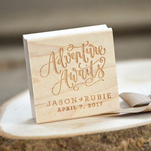 Puede incluir: Sello cuadrado de madera con el texto "Adventure Awaits" en escritura cursiva. Debajo del texto se lee "Jason &hearts; Rubie April 7, 2017".