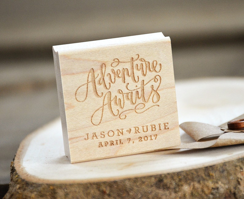 Puede incluir: Sello cuadrado de madera con el texto "Adventure Awaits" en escritura cursiva. Debajo del texto se lee "Jason &hearts; Rubie April 7, 2017".