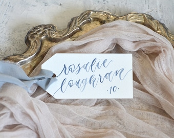 May include: A white tag with the name "rosalie loughran" in blue calligraphy and the number "10". The tag is tied with a blue ribbon and rests on a beige fabric, with a gold-coloured decorative tray in the background.