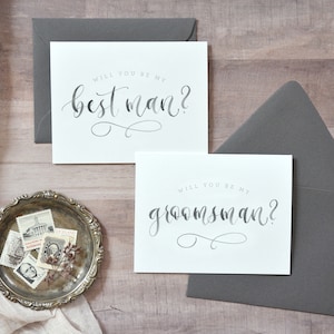 May include: Two white cards with gray calligraphy text. The top card says "Will you be my best man?" and the bottom card says "Will you be my groomsman?"