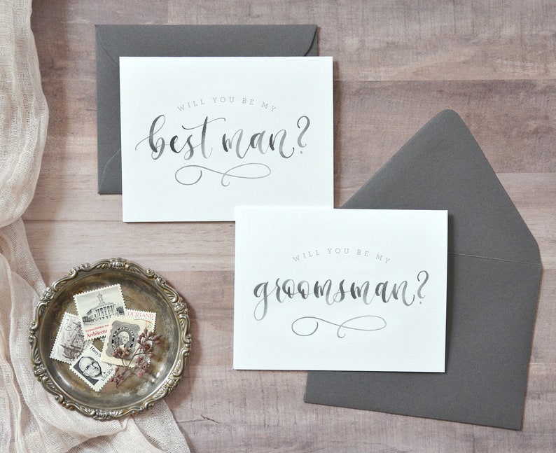 May include: Two white cards with gray calligraphy text. The top card says "Will you be my best man?" and the bottom card says "Will you be my groomsman?"