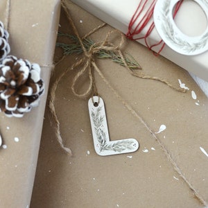 May include: A brown paper gift bag with a white letter L gift tag tied with twine. The gift tag has a green and white floral design. The bag is decorated with white paint splatters and pine cones.