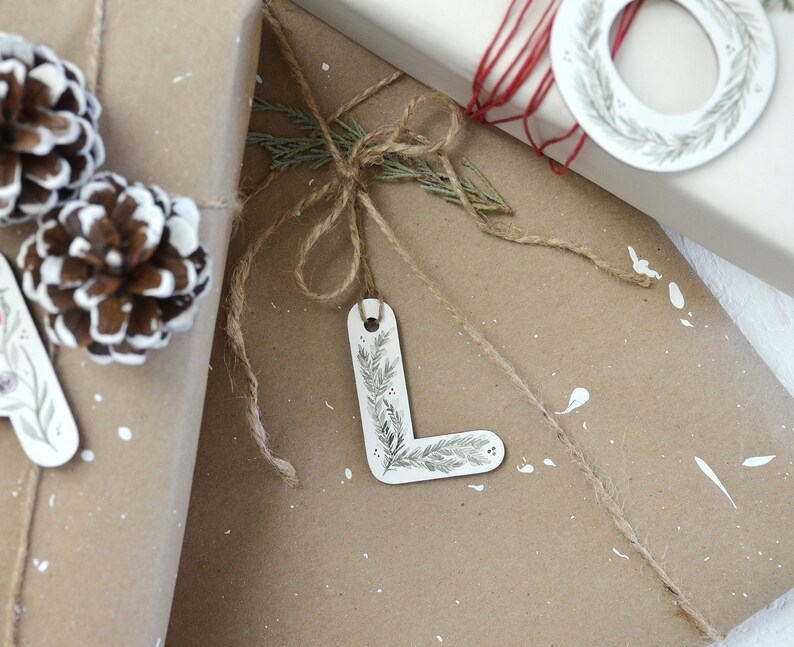 May include: A brown paper gift bag with a white letter L gift tag tied with twine. The gift tag has a green and white floral design. The bag is decorated with white paint splatters and pine cones.