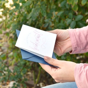 May include: A white card with a blue envelope. The card has pink calligraphy that says "Will you be my bridesmaid?"