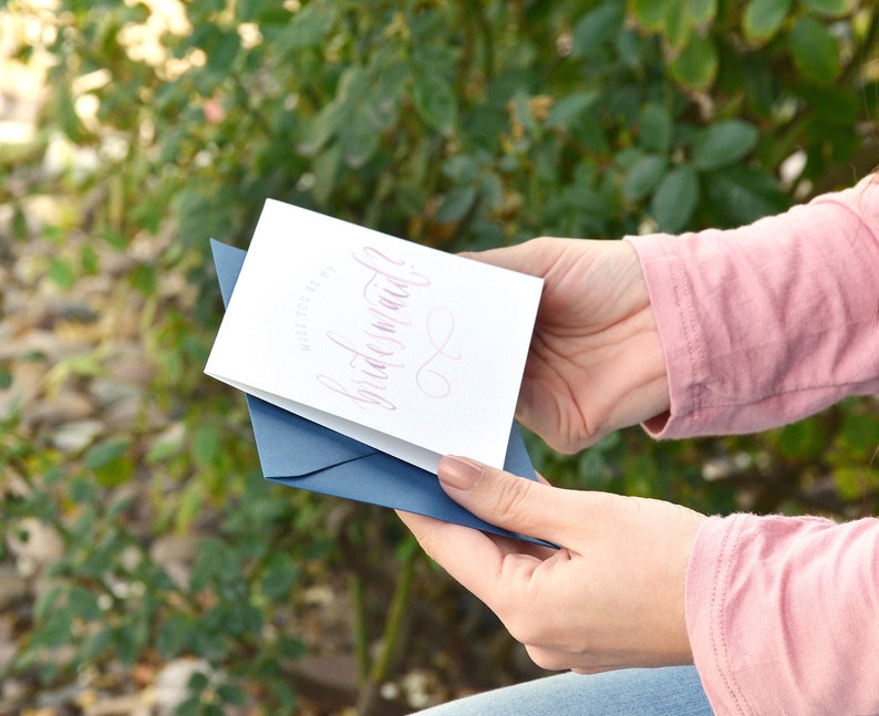 May include: A white card with a blue envelope. The card has pink calligraphy that says "Will you be my bridesmaid?"
