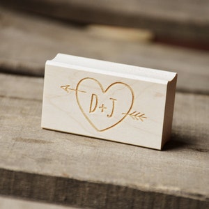 Può includere: Timbro in legno con un design a forma di cuore e una freccia che lo attraversa. Il cuore contiene le iniziali "D+J" all'interno.