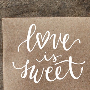 May include: A brown paper bag with the phrase "love is sweet" written in white cursive lettering with a heart shape above the word "love".