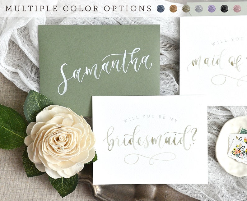 May include: A white card with the text "Will you be my bridesmaid?" written in silver script. The card is on a green envelope with the name "Samantha" written in silver script.