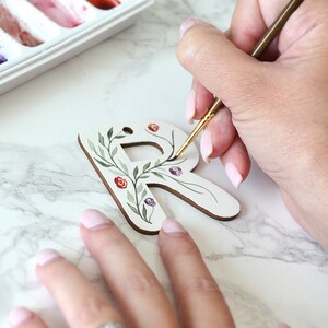 May include: A person paints a wooden letter R with a floral design. The letter is white and the flowers are painted in shades of green, red, and purple.