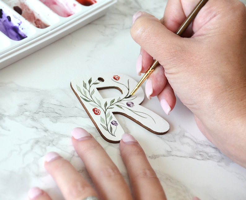 May include: A person paints a wooden letter R with a floral design. The letter is white and the flowers are painted in shades of green, red, and purple.