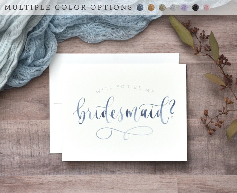 May include: White card with the text "Will you be my bridesmaid?" in blue calligraphy. The card is on a wooden surface with a blue fabric and a sprig of greenery.