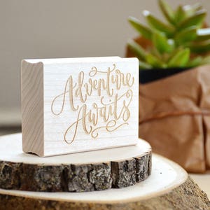Puede incluir: Un sello de madera con el texto "Adventure Awaits" en una fuente cursiva. El sello est&aacute; sobre una superficie de madera con una peque&ntilde;a planta en maceta en el fondo.