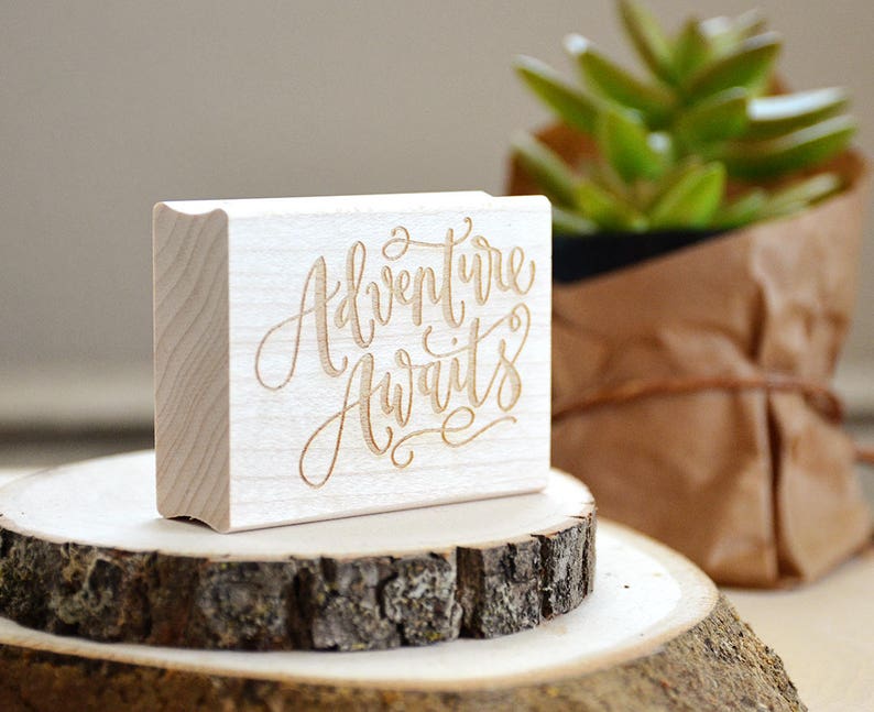 Puede incluir: Un sello de madera con el texto "Adventure Awaits" en una fuente cursiva. El sello est&aacute; sobre una superficie de madera con una peque&ntilde;a planta en maceta en el fondo.