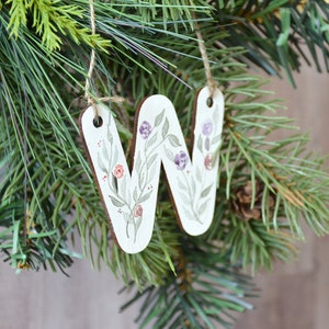 May include: A wooden ornament shaped like the letter W, painted with a floral design in shades of green, purple, and pink. The ornament is hanging from a twine string.