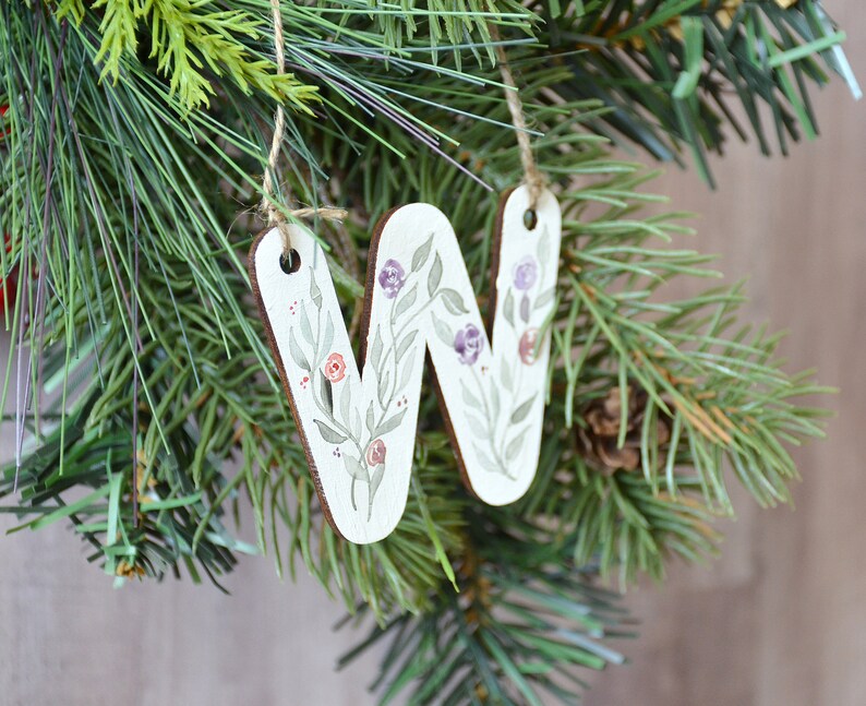May include: A wooden ornament shaped like the letter W, painted with a floral design in shades of green, purple, and pink. The ornament is hanging from a twine string.