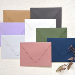 May include: A set of ten envelopes in various colors, including lavender, white, light brown, dark gray, green, and navy blue. The envelopes are arranged in a staggered pattern on a white textured surface.