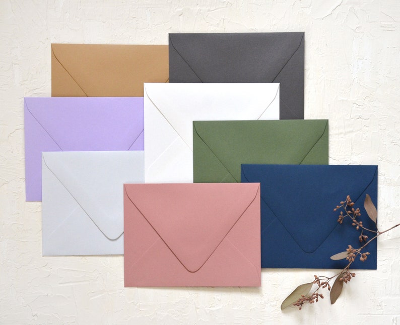 May include: A set of ten envelopes in various colors, including lavender, white, light brown, dark gray, green, and navy blue. The envelopes are arranged in a staggered pattern on a white textured surface.