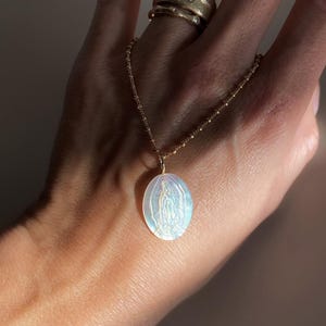 Op de afbeelding: Een goudkleurige ketting met een ovale hanger met een afbeelding van de Maagd Maria. De hanger heeft een parelmoer, iriserende afwerking. De ketting wordt door een hand vastgehouden en er zijn twee gouden ringen op de vinger zichtbaar. De hanger is ongeveer 2,5 cm lang.