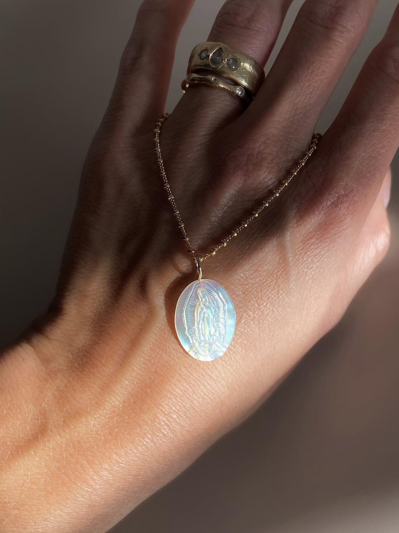 Op de afbeelding: Een goudkleurige ketting met een ovale hanger met een afbeelding van de Maagd Maria. De hanger heeft een parelmoer, iriserende afwerking. De ketting wordt door een hand vastgehouden en er zijn twee gouden ringen op de vinger zichtbaar. De hanger is ongeveer 2,5 cm lang.