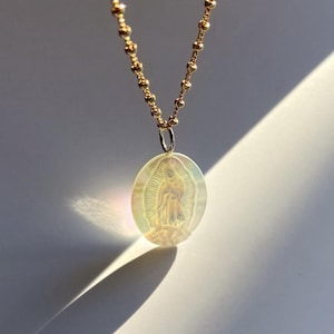 Op de afbeelding: Een goudkleurige ketting met een ovale hanger met een afbeelding van de Maagd Maria. De hanger heeft een iriserende, parelmoerachtige afwerking. De ketting is gemaakt van kleine, ronde kralen.