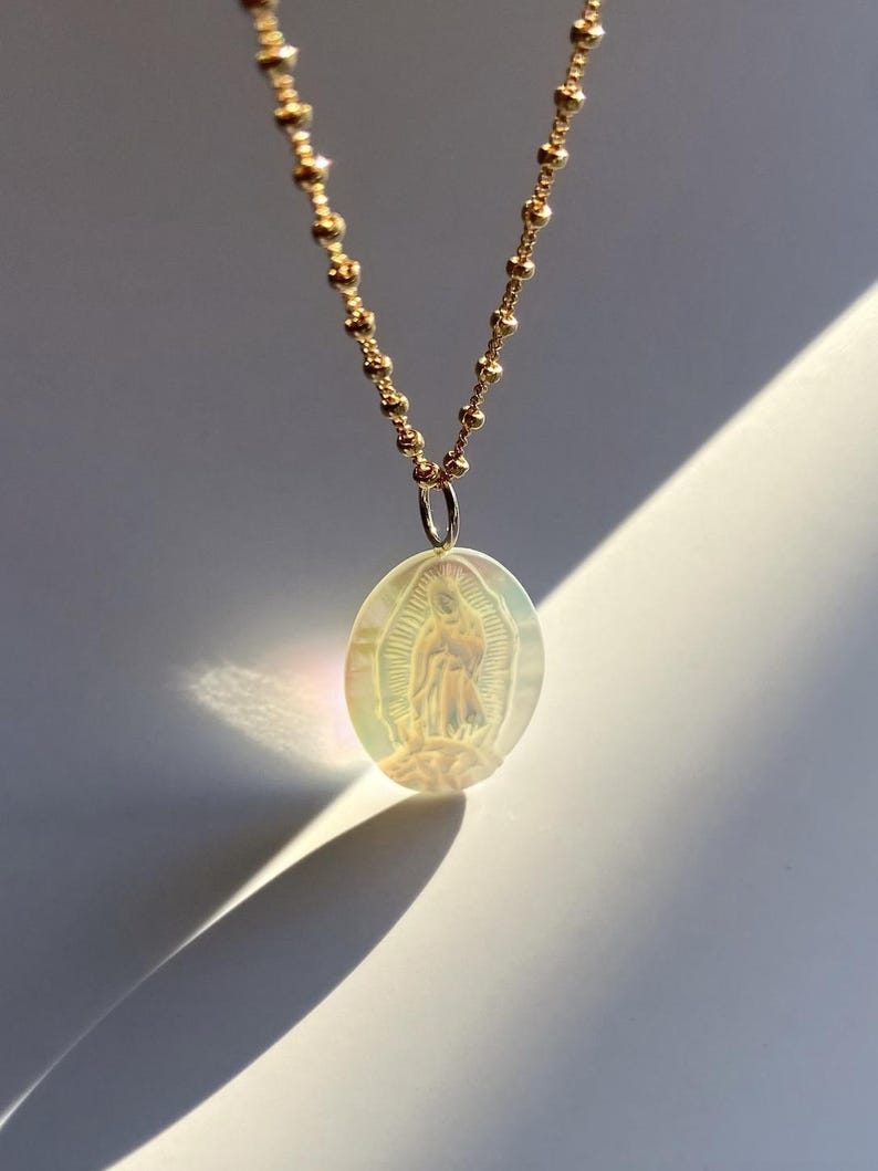 Op de afbeelding: Een goudkleurige ketting met een ovale hanger met een afbeelding van de Maagd Maria. De hanger heeft een iriserende, parelmoerachtige afwerking. De ketting is gemaakt van kleine, ronde kralen.