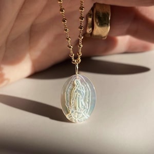 Op de afbeelding: Gouden ketting met een ovale hanger met een religieuze figuur. De hanger heeft een iriserende glans en gedetailleerde gravure. De ketting is gemaakt van kleine, ronde gouden kralen. Religieuze sieraden.