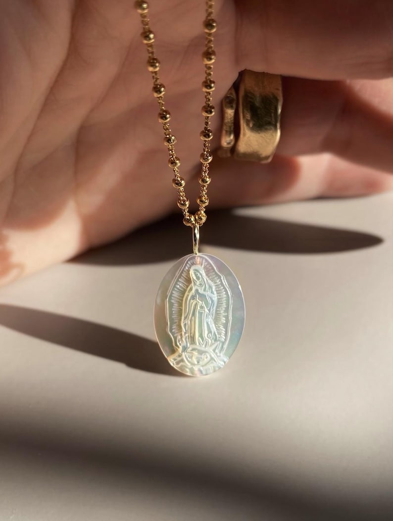 Op de afbeelding: Gouden ketting met een ovale hanger met een religieuze figuur. De hanger heeft een iriserende glans en gedetailleerde gravure. De ketting is gemaakt van kleine, ronde gouden kralen. Religieuze sieraden.