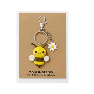 May include: A yellow and black bee keychain with a daisy charm, attached to a silver metal clasp and keyring. The keychain is on a brown card with the text "YoursNotably Fun & Cheerful keychains".