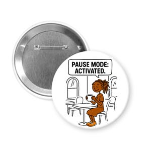 May include: A white button pin with a cartoon illustration of a person sitting at a table, holding a mug. The text "PAUSE MODE: ACTIVATED." is in a speech bubble. A second, unadorned pin is also visible.