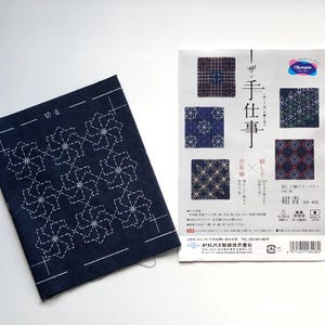May include: A Sashiko embroidery kit featuring a dark blue fabric panel with a white geometric star pattern, an instructional paper guide with Japanese text and illustrations, a bundle of multi-colored embroidery thread, and a small needle pack.