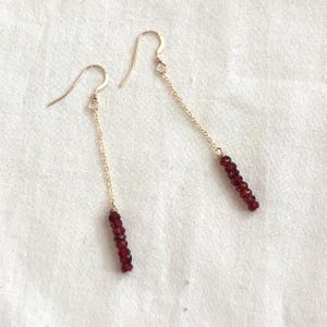 May include: A pair of gold-colored earrings with a chain and a cluster of small, dark red beads. The earrings hang from a curved hook. The beads are approximately 0.25 inches in length.