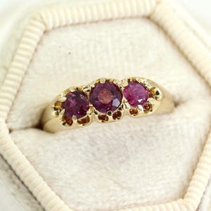 May include: A gold ring featuring three round, deep pink gemstones. The ring is set in a classic style, with the gemstones slightly raised and surrounded by small decorative elements. The ring is presented in a white, hexagonal jewellery box.