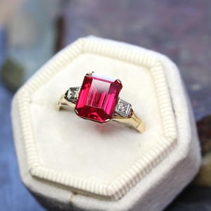 Può includere: Un anello d'oro con una grande gemma rossa brillante di forma rettangolare. Due gemme chiare, quadrate e più piccole affiancano la pietra principale. L'anello è presentato in un cofanetto di velluto bianco.