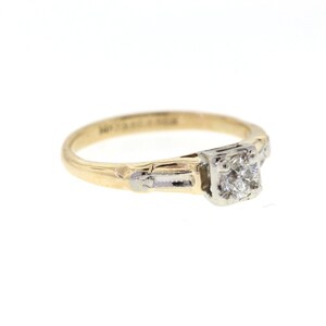 May include: A gold and white engagement ring with a single round diamond in the center. The ring has a delicate design with a band that is slightly wider at the center.