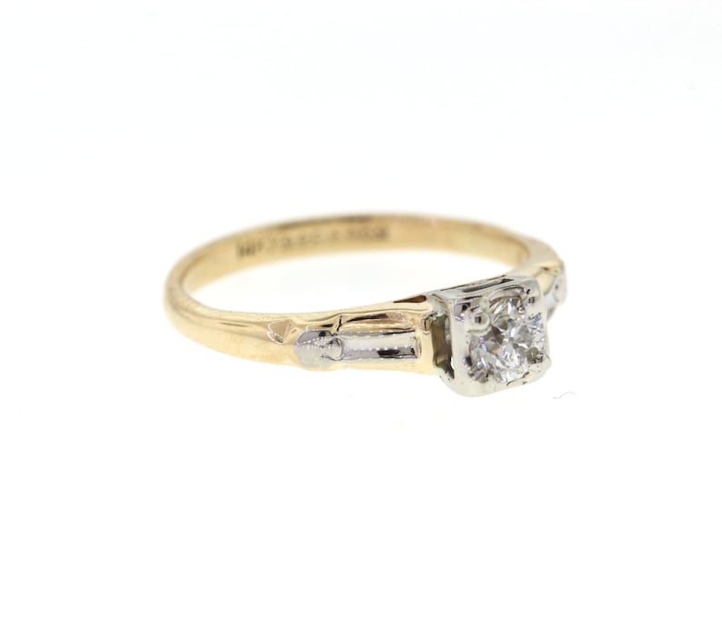May include: A gold and white engagement ring with a single round diamond in the center. The ring has a delicate design with a band that is slightly wider at the center.