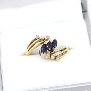 May include: A gold ring with a unique design, featuring three dark blue marquise-cut gemstones and small, round, clear gemstones. The ring is presented in a gray jewelry box with a white interior, showcasing its elegant details.