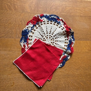 May include: A round, white crocheted doily with a scalloped border featuring red and blue yarn accents, layered beneath a square, solid red fabric napkin with a thin white border trim along the edges, creating a layered textile arrangement.