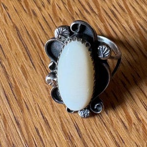 May include: A silver ring with an oval, off-white stone. The ring has a decorative design with leaf-like accents around the stone. The ring is set against a wooden surface.