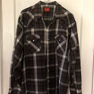 May include: A long-sleeved plaid shirt in shades of brown, black, and white hangs on a metal hanger. The shirt has two chest pockets with silver snap closures and a collar. The brand name "PLAINS" is visible on a red tag.