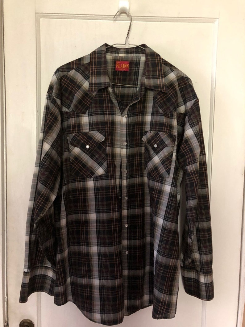 May include: A long-sleeved plaid shirt in shades of brown, black, and white hangs on a metal hanger. The shirt has two chest pockets with silver snap closures and a collar. The brand name "PLAINS" is visible on a red tag.