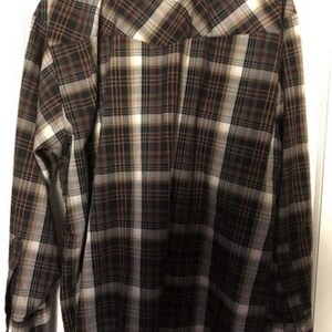 May include: A long-sleeved plaid shirt in shades of brown, black, and white. The shirt has a button-down front, button cuffs, and a classic collar. The back of the shirt is visible, hanging against a white door.