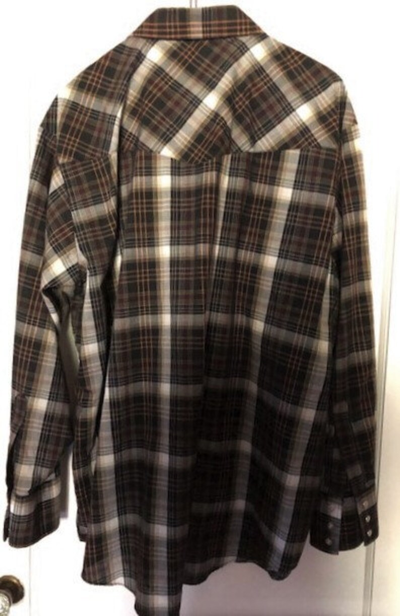 May include: A long-sleeved plaid shirt in shades of brown, black, and white. The shirt has a button-down front, button cuffs, and a classic collar. The back of the shirt is visible, hanging against a white door.